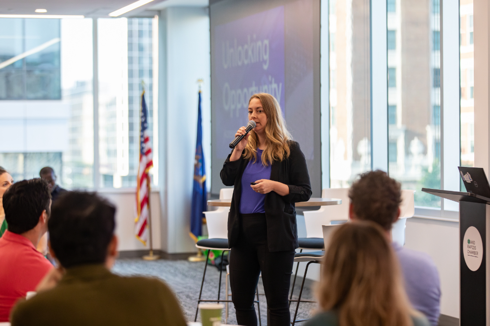 Rachel Salinas with microphone standing to speak to audience at tables. Projector screen reads Unlocking Potential and there are US and Michigan flags standing at left