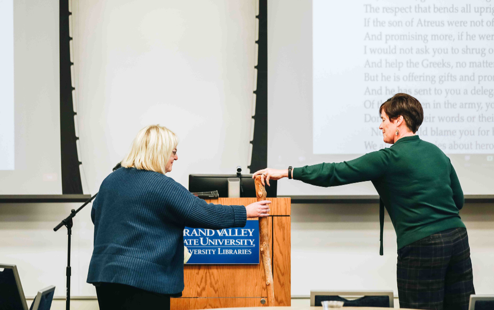 two women pass a wooden scepter between them, a projection screen in the background