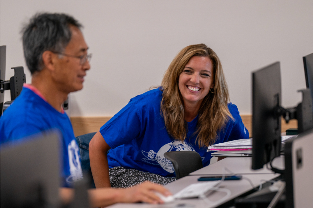 two people in blue t-shirts in computer lab, woman at right is smiling facing the camera