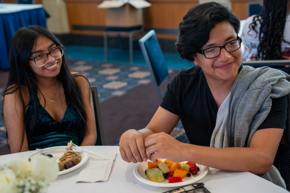 two students seated at table smiling, plates of food in front of them