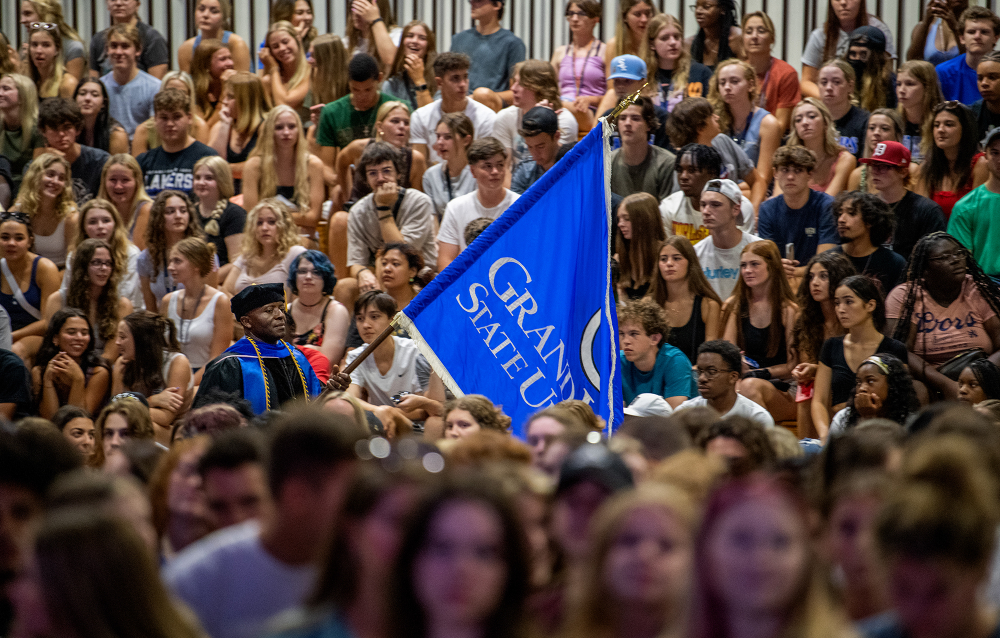 man in academic regalia carries GVSU flag through crowd of students seated on bleachers and chairs in the Fieldhouse