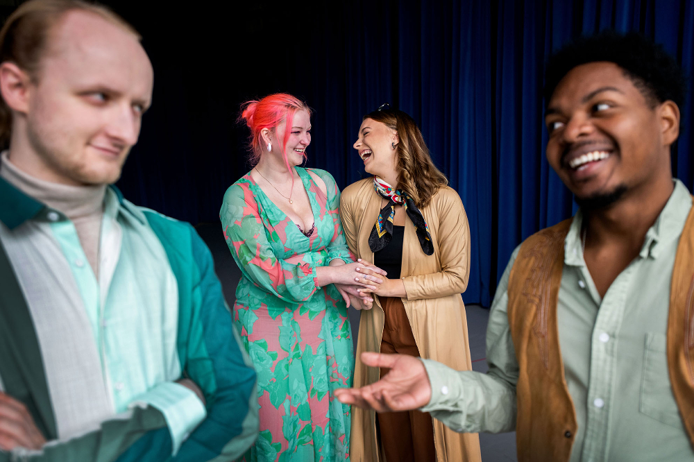 four actors in costumes, two women at back holding hands, two men in front glaring at each other. Man on right has big smile on his face, gesturing with his right hand