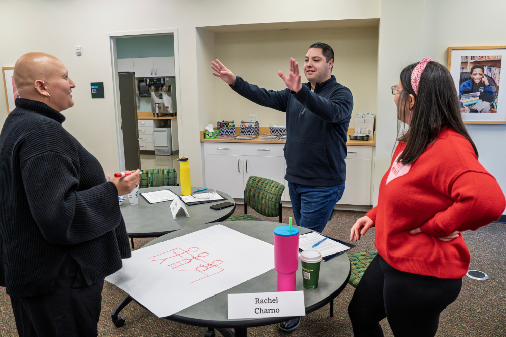 three people standing around round table filled with papers, markers and tumblers