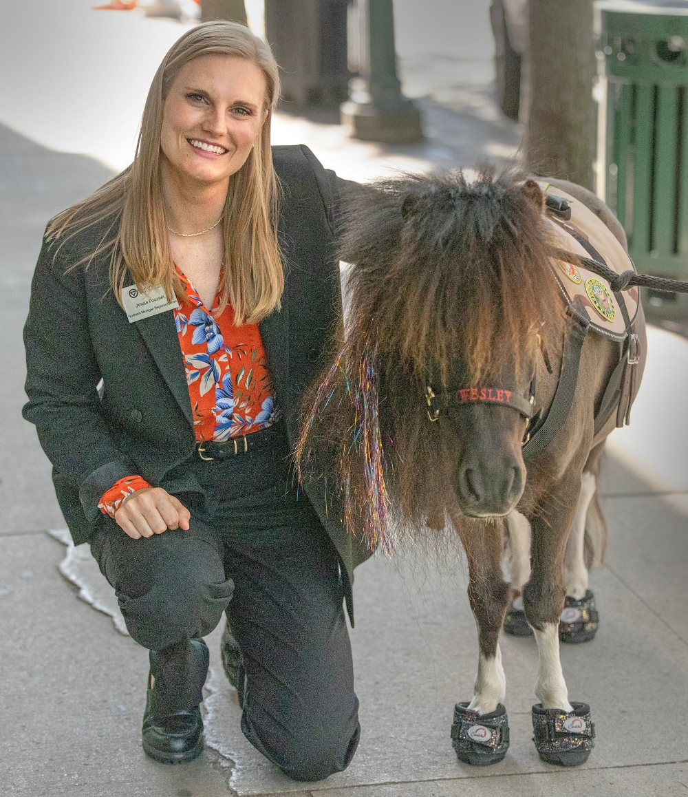Jessie Fousset kneels on a sidewalk next to a small therapy horse
