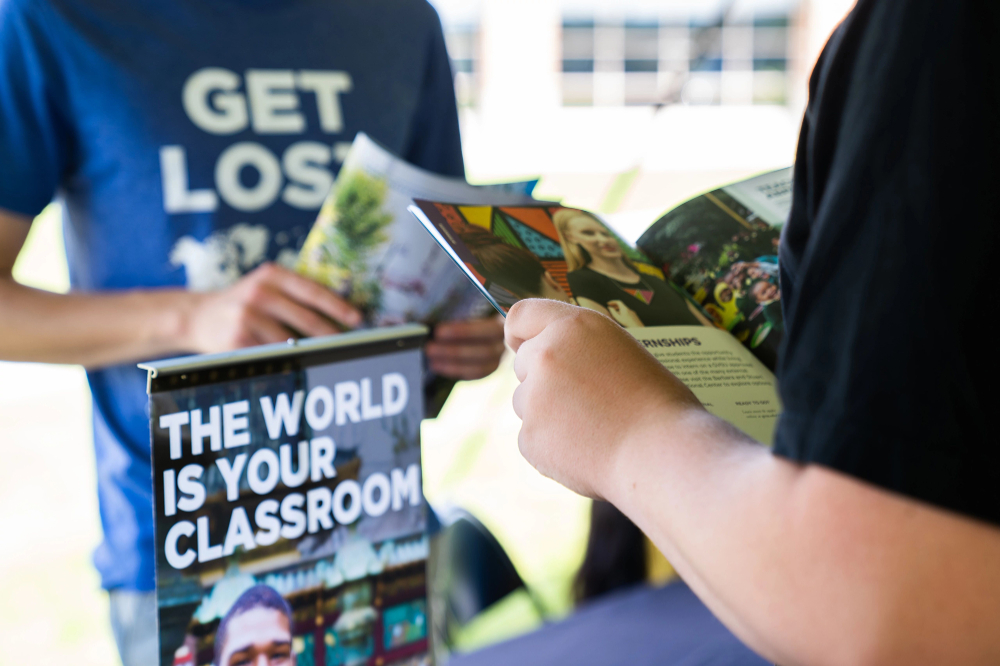 two people one standing, one sitting reading magazines, a poster nearby reads "The World Is Your Classroom" One person's t-shirt says Get Lost