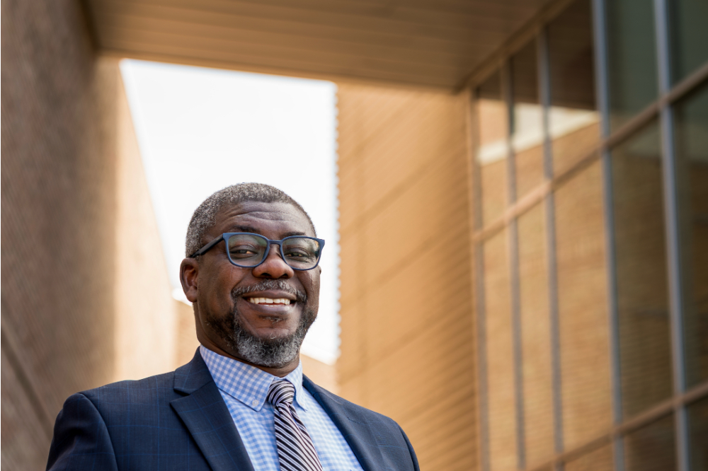 Adebayo "Bayo" Ogundipe in blue suit and checked light blue shirt standing outside the Kennedy Hall with sunlight behind him