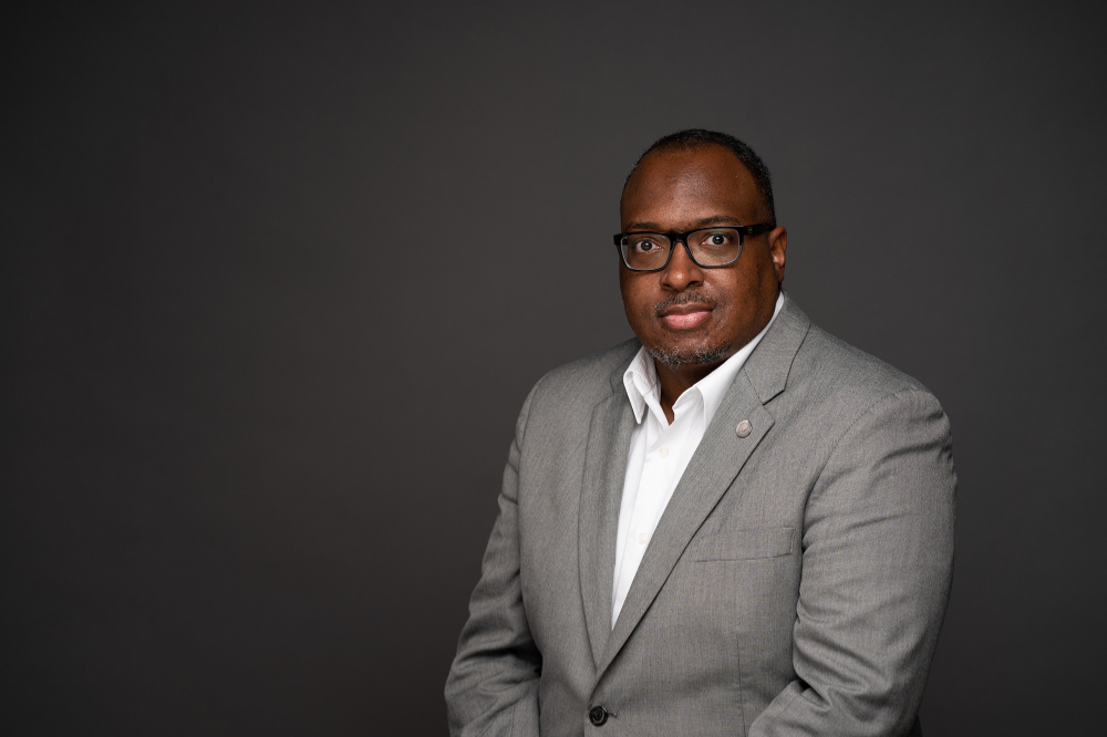 portrait of Dwayne Tunstall, seated in grey suit jacket with white shirt and no tie