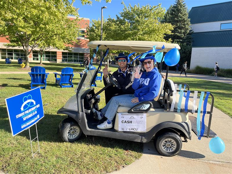 two people in a golf cart that is decorated with balloons and streamers, a sign for Student Philanthropy Week is at left in the ground