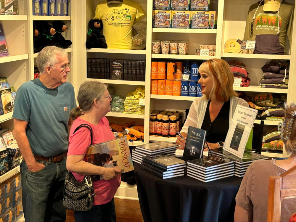 at a bookstore, a woman holds up her book at a signing while two people stand in line to chat