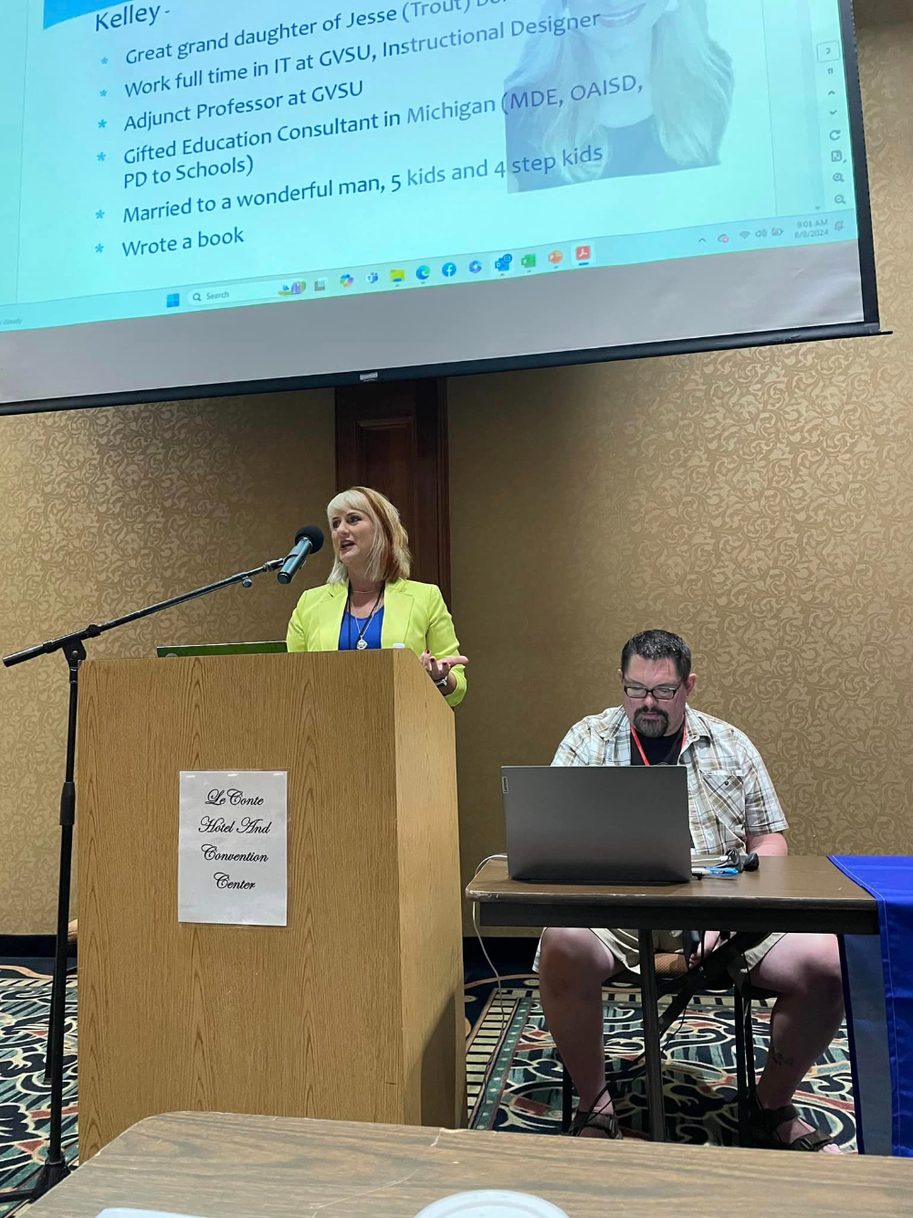 Kelley Senkowski gives a presentation behind a podium and microphone, man seated at table to her right with open laptop