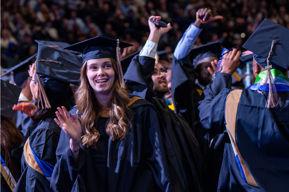 students in black graduation robes and caps stand, smile and look to their supporters in the audience