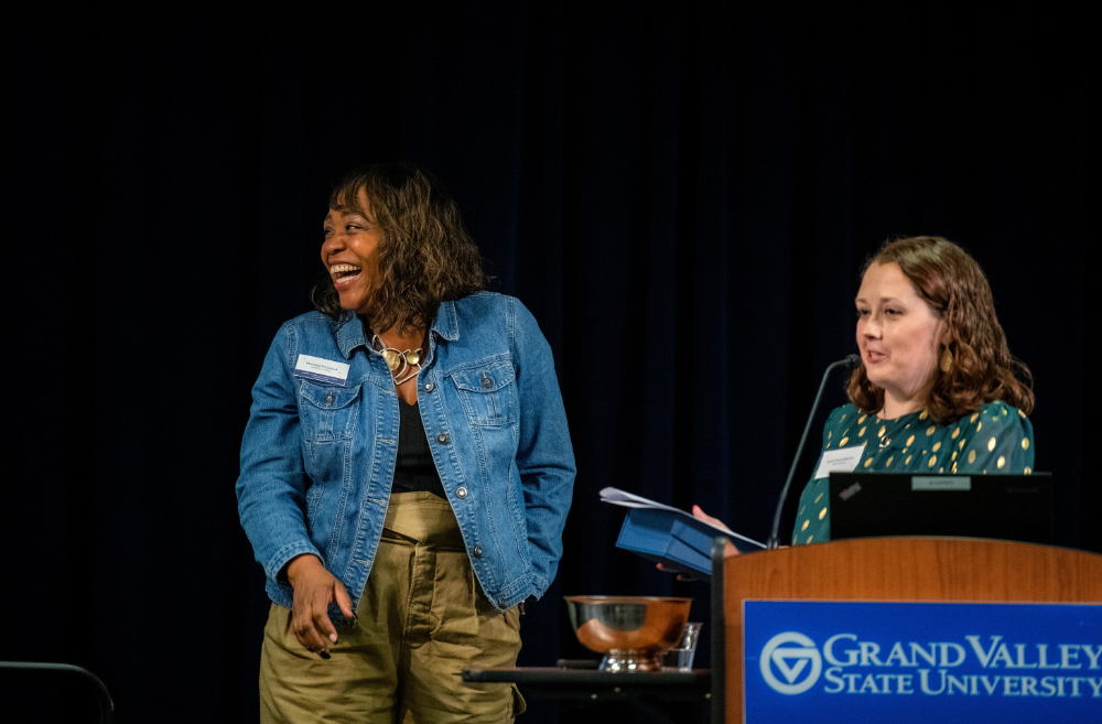 two women on stage, woman on left laughing while woman behind podium presents her with an award