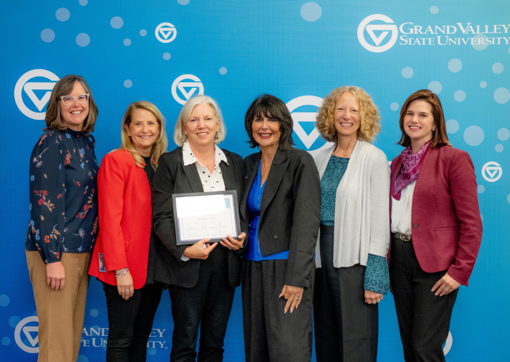 six women standing against a GVSU backdrop; woman third from left holds a certificate