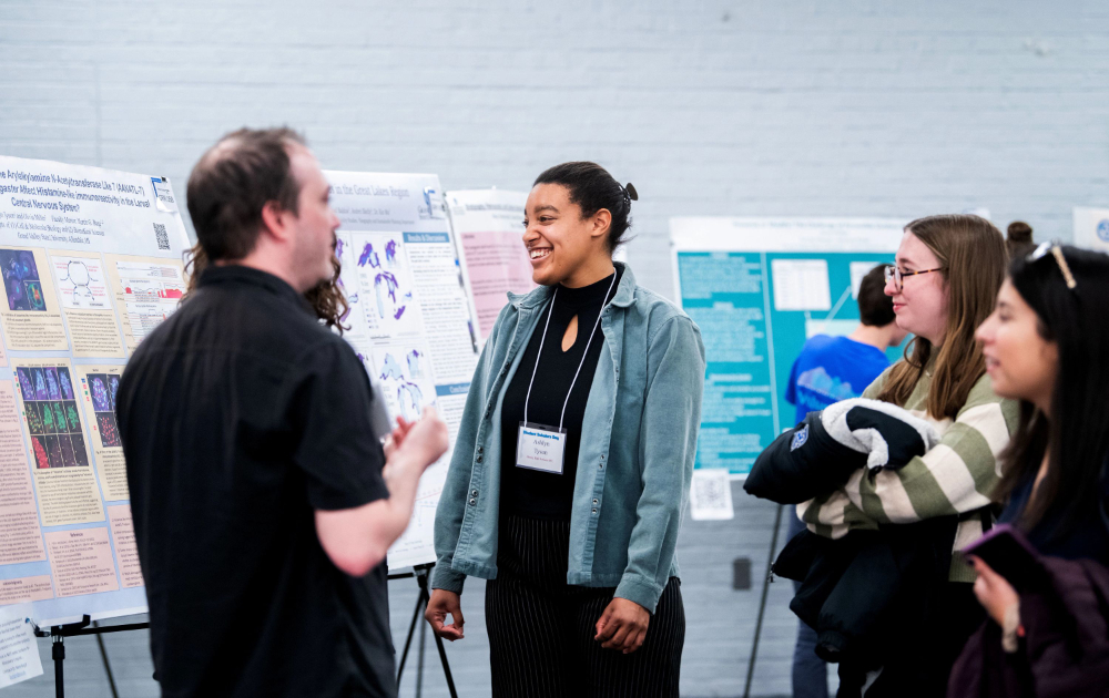 posters lining the wall on easels; a student presenter talks with someone with his back to the camera, two others are at right