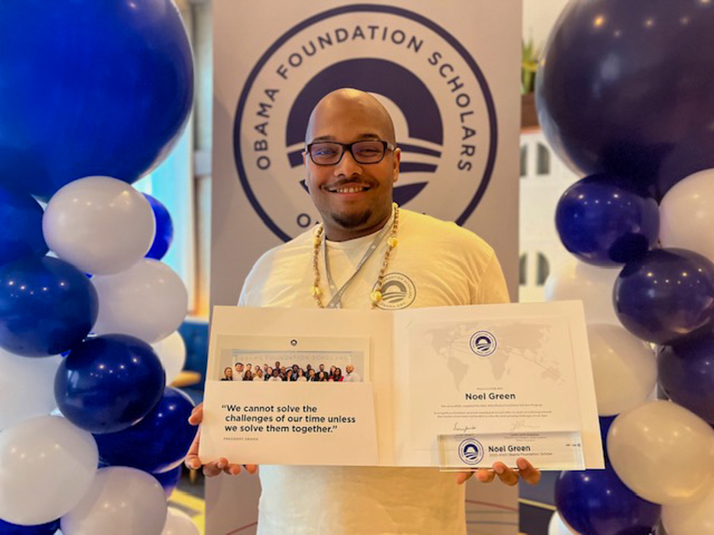 man holds certificate folder in front of background of white and blue balloons and the Obama Foundation Scholars program vertical banner