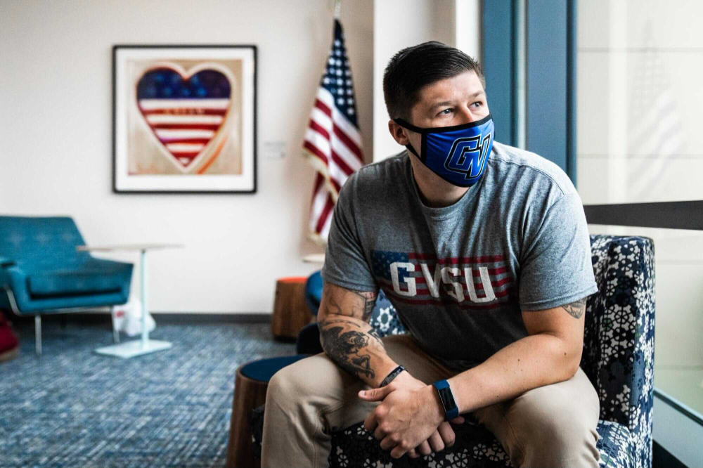 student veteran sits in chair in veterans lounge, behind him is American flag and frame on wall with heart decorated similar to flag, with stripes and stars