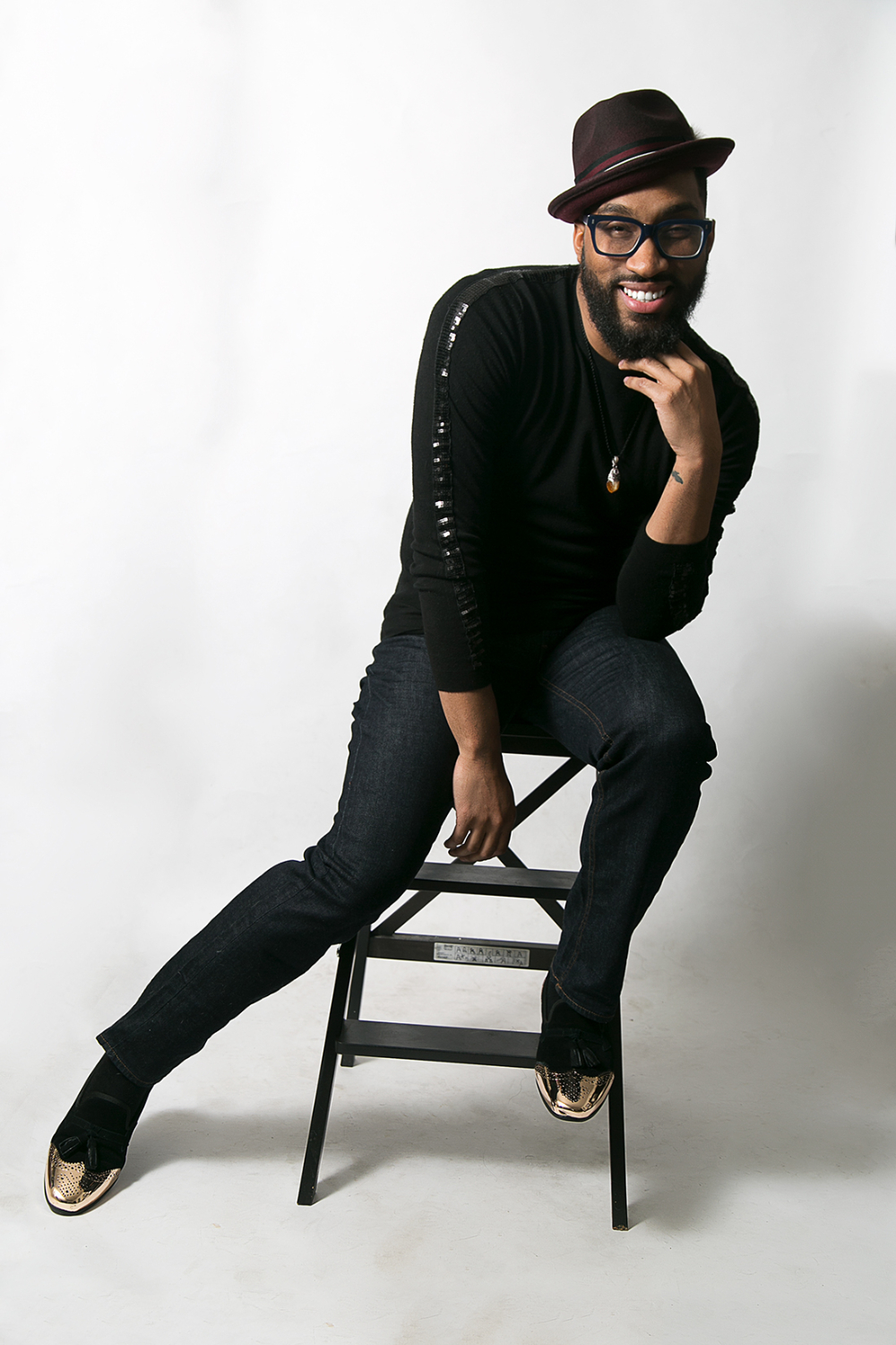 Edgar Page is seated on a chair, leaning to the left, he is wearing a black hat and glasses, with a black outfit.