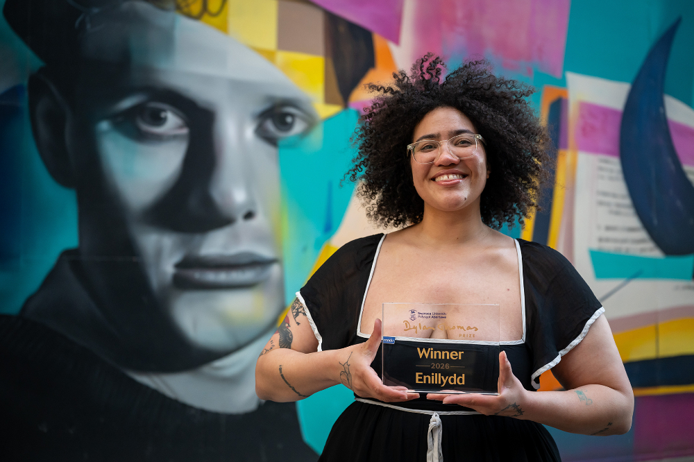 Sasha Debevec-McKenney holds a glass award in front of a colorful mural of Dylan Thomas