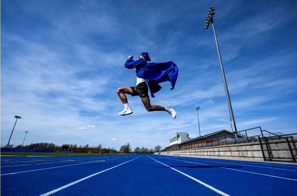 Marcus Gelpi in a blue commencement gown and cap jumps over the track at Lubbers Stadium
