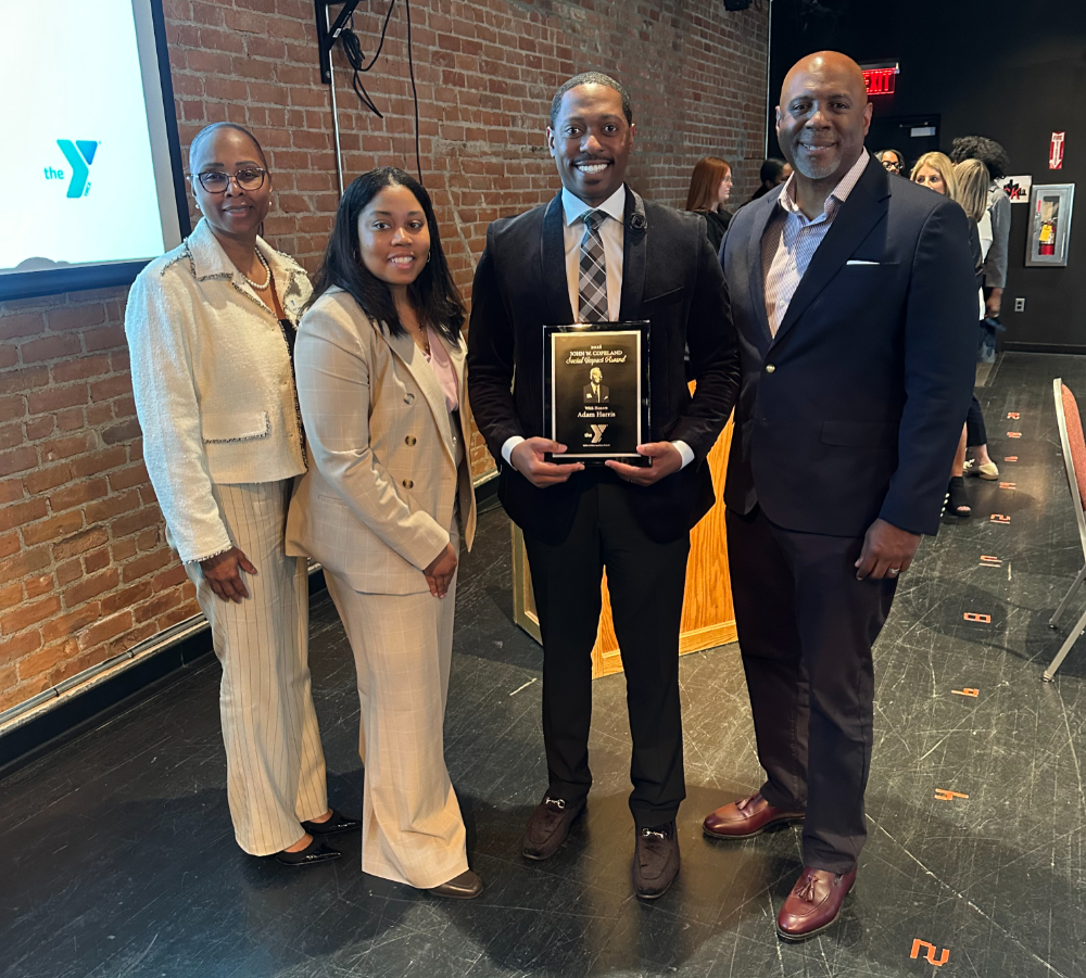 Adam Harris, second from right, holds an award. Two women in light colored suits stand at Harris's left and a man in a suit jacket without a tie is at right.