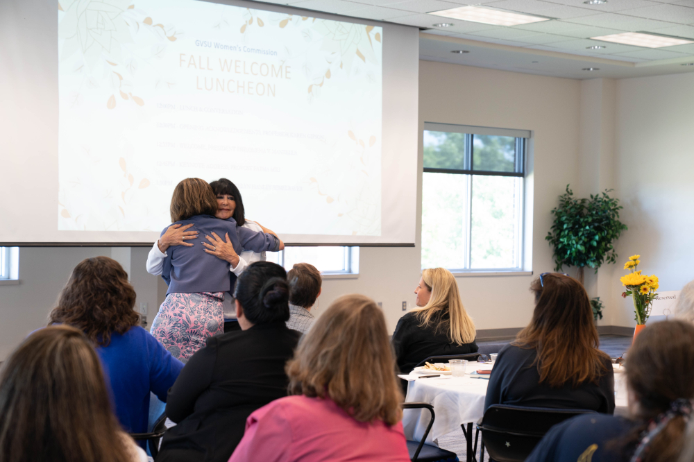President Mantella and Provost Mili share an embrace at the front of a room, women seated at tables look on