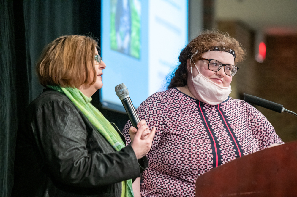 two people at podium, person on left with microphone; person at right is wearing a face mask; projector screen down and lit up in the background