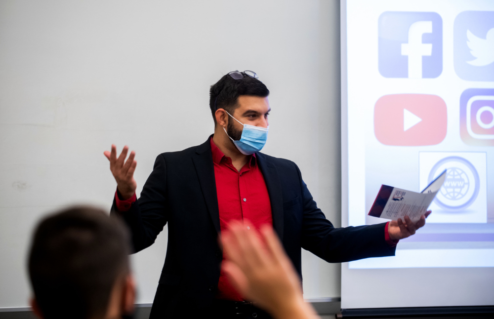 person in sports jacket and red shirt stands and gestures at front of room, holding paperwork