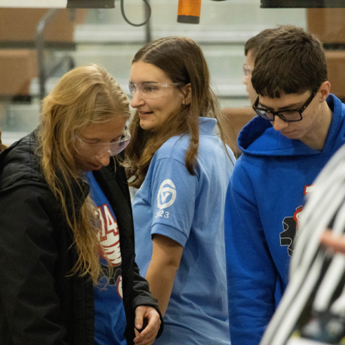 Madelyn Rynsburger in blue golf shirt wears safety goggles at a FIRST Robotics event