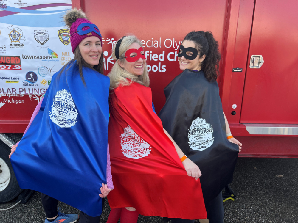 three people standing with the backs to camera in capes (blue, red and black) and eye masks