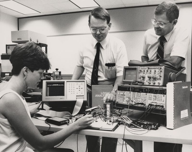 black and white photo in the 1980s of a student in front of computer equipment with two faculty standing looking on