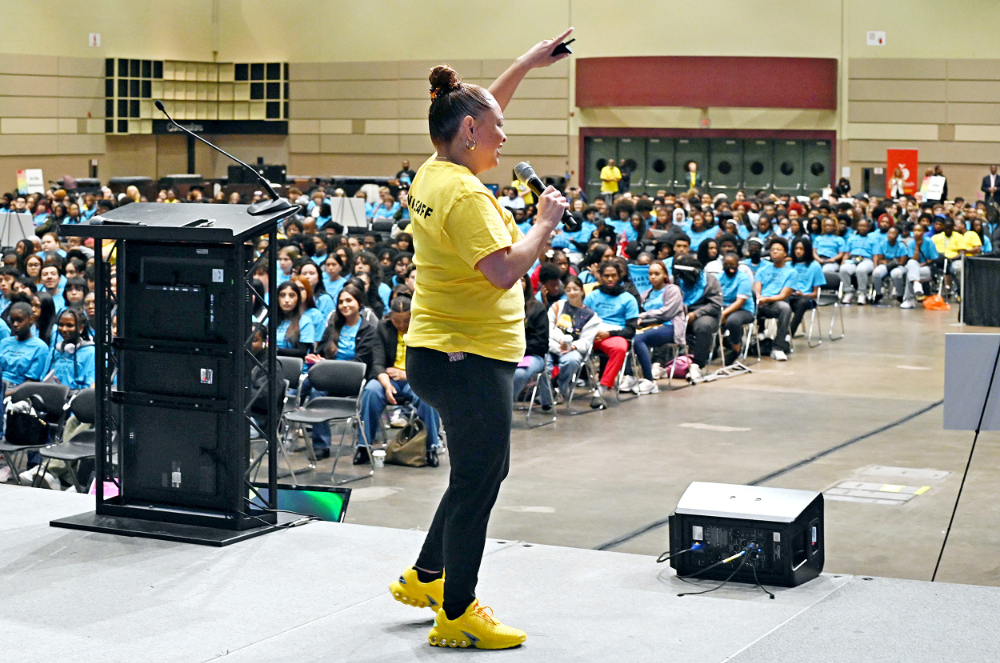 woman on stage in yellow t-shirt, hundreds of students in blue t-shirts seated in rows in large room