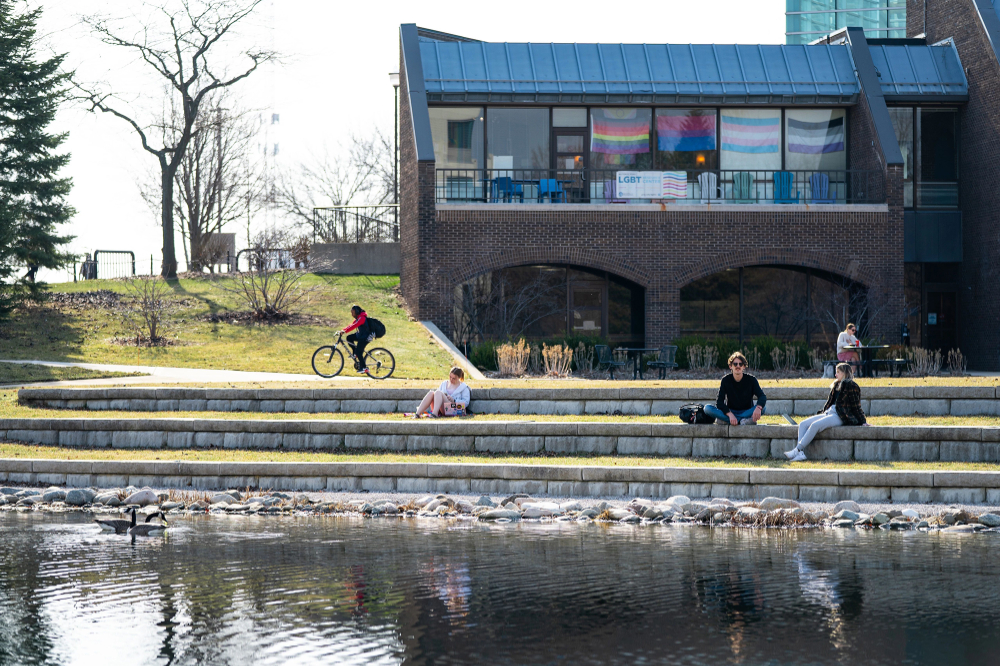 students sitting on ledges near Zumberge Pond, one bicyclist in background; pride flags in background on Kirkhof Center office