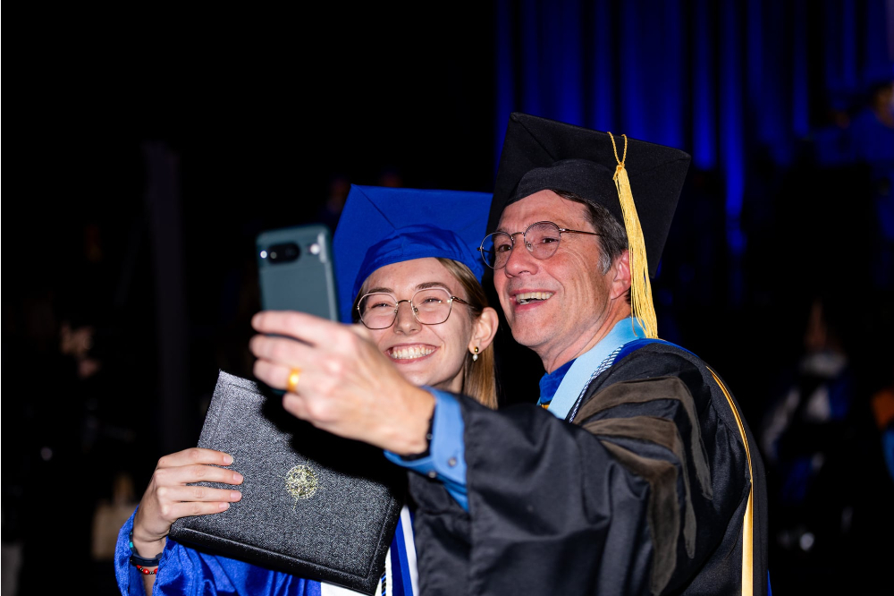 faculty member on right next to a new graduate taking a selfie during the December commencement at Van Andel Arena
