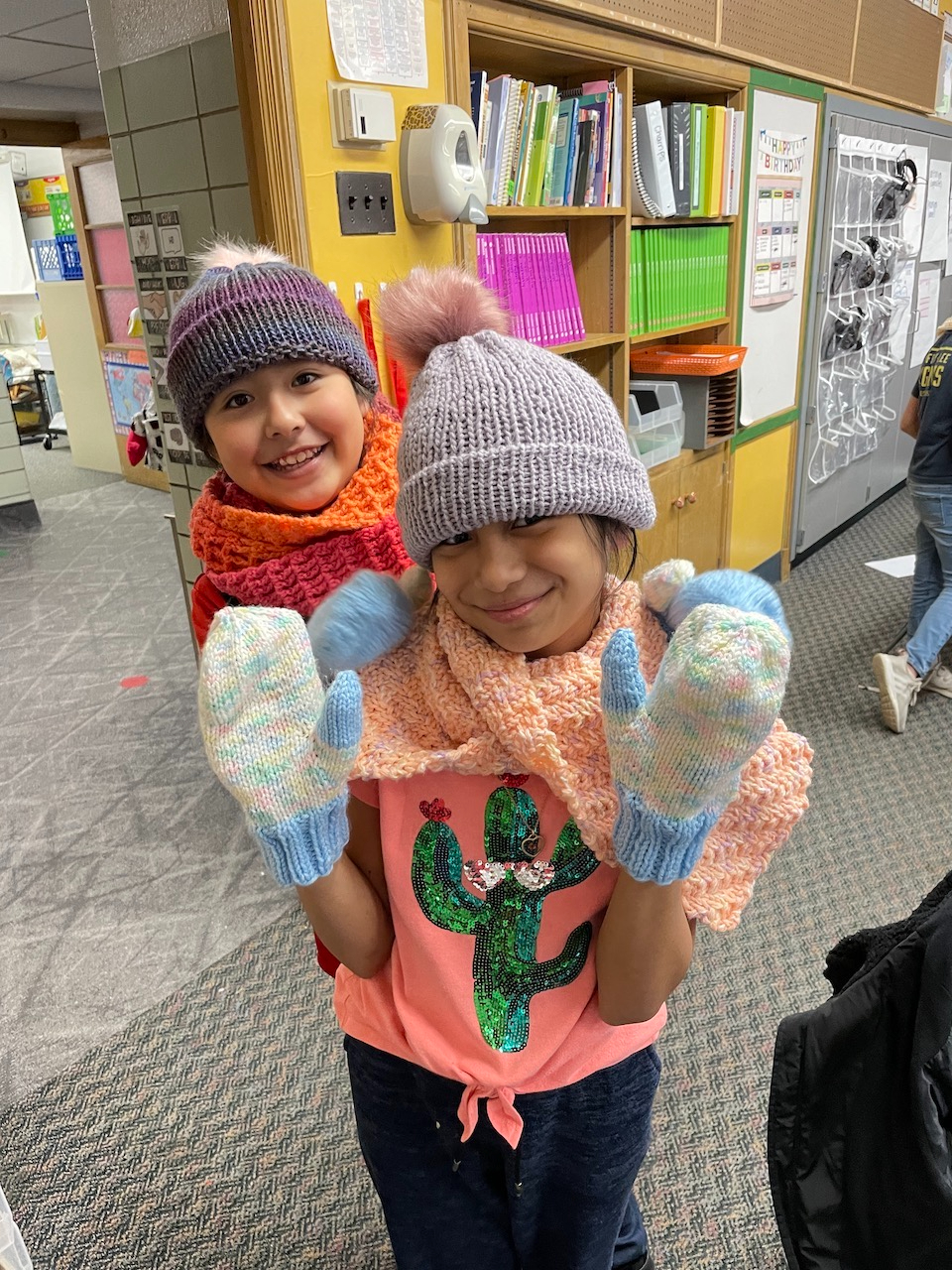 elementary students show off their knitted mittens and hats inside a classroom