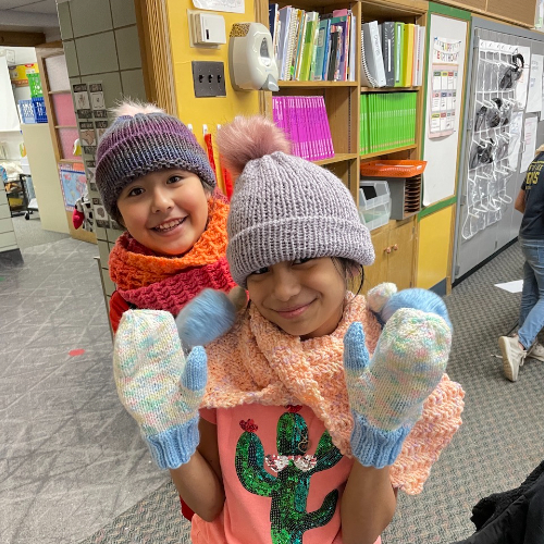 elementary students show off their knitted mittens and hats inside a classroom