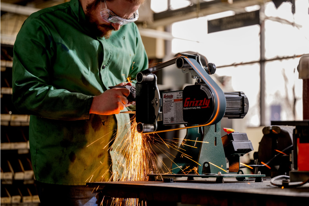 student in green safety lab coat and goggles using a grinder to shape a tool, sparks flying