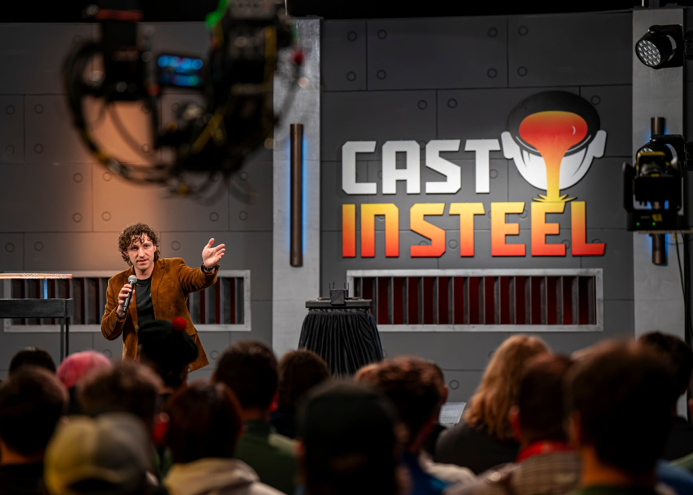 man in brown coat gestures to people seated in chairs, Cast in Steel sign to his right