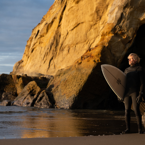 Ben Moon holds a surfboard on the Oregon coast in front of large rock formation