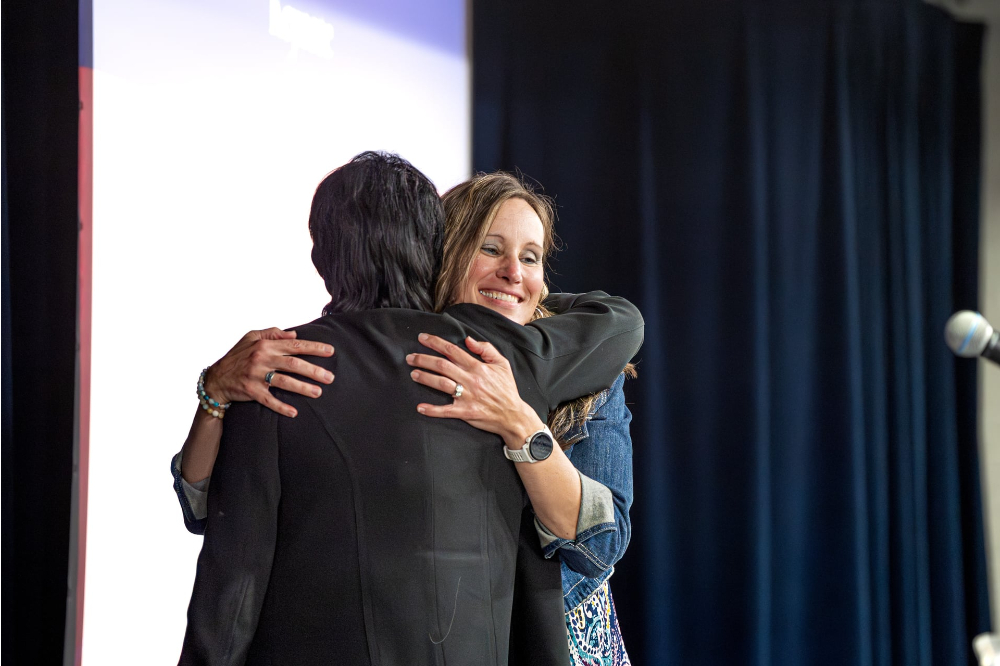 President Mantella in black blazer hugs Erica Hamilton on a stage as she gets ready to speak