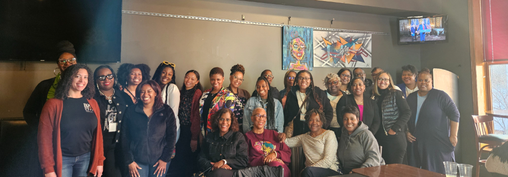 25 Black women in a restaurant, four are seated, rest are standing in two staggered rows