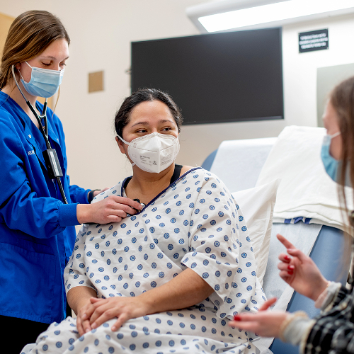 in a hospital-like room with patient bed, a nursing student uses a stethoscope to listen to a standardized patient in a hospital gown, while a student seated in a chair uses hand gestures while listening and interpreting