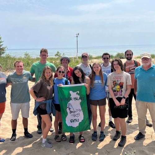 group of students and faculty standing on beach in front of lake and holding GVSU Beaver Island green flag