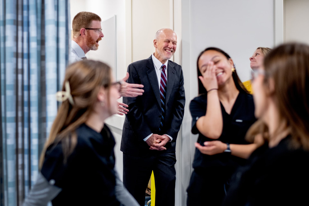 Jeffrey Potteiger leans against wall laughing with colleagues, while students in black scrubs are in the forefront.