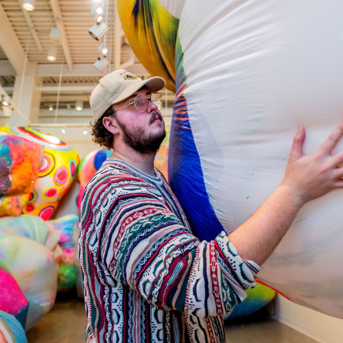 male student installs a large piece of artwork, with colorful pieces stacked behind him in a gallery