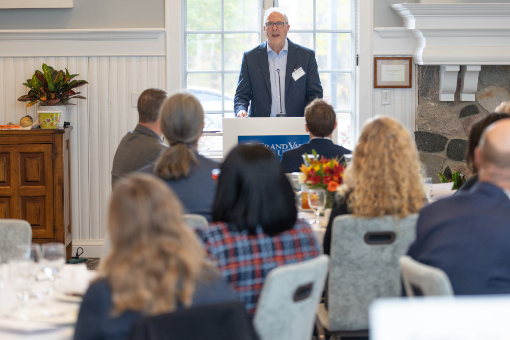 Peter Riemersma stands at podium inside alumni house, people seated at round tables