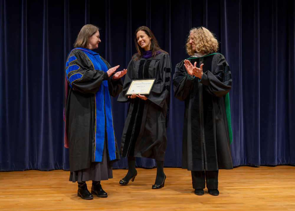 three people standing on stage, woman in center holds framed certificate