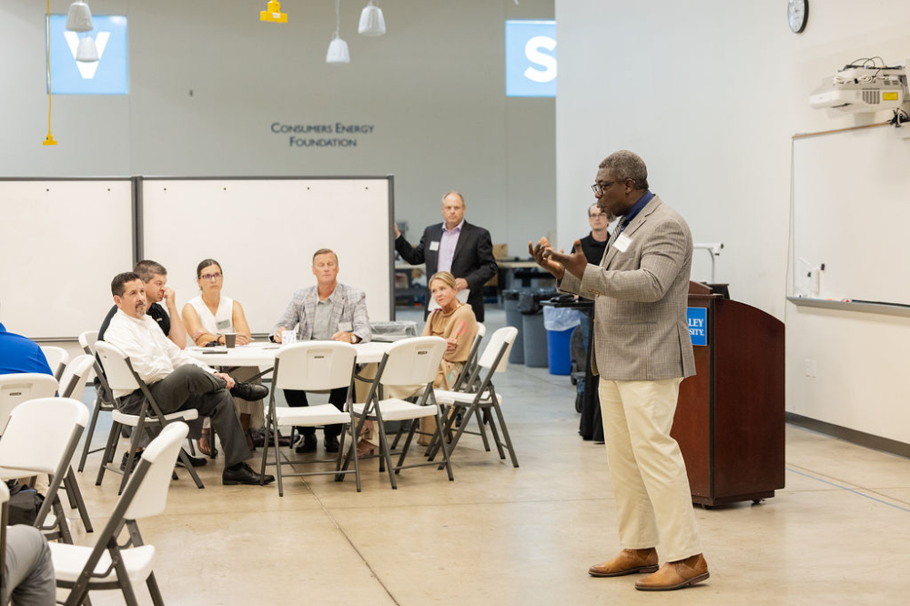 Bayo Ogundipe in suit with microphone in front of room with round tables and people seated