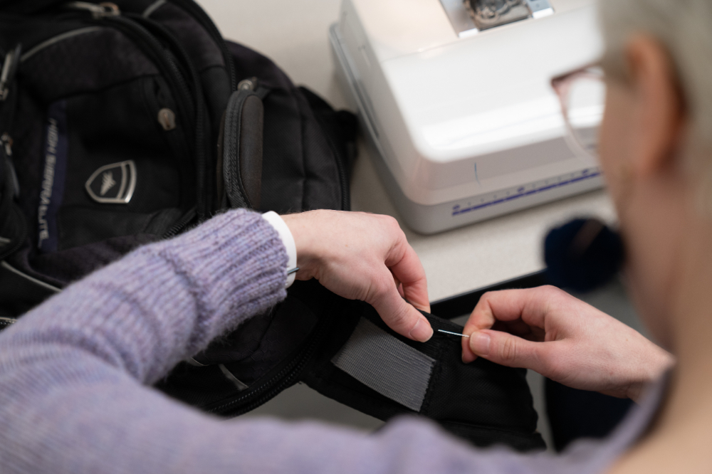 person sewing a repair to a backpack strap, a sewing machine base is on the table