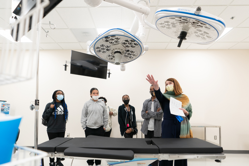 students and an adult stand in front of an operating table looking at large ceiling lights in the Simulation Center
