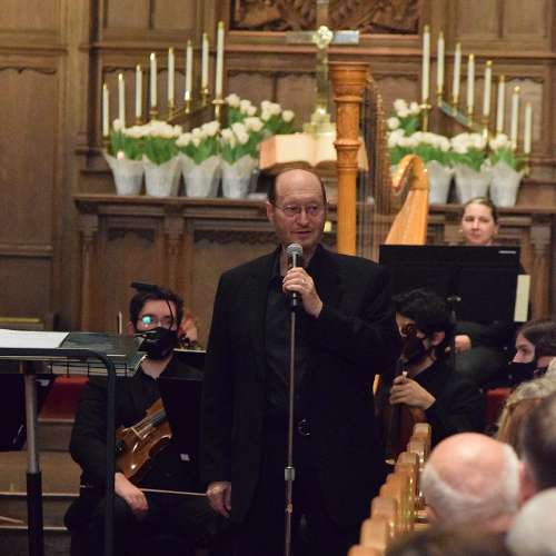 Henry Duitman speaks into a microphone. Musicians seated behind him and an alter filled with candles and flowers
